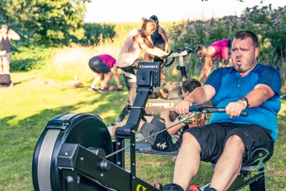 Mann beim Rudern im Freien, umgeben von anderen Sportlern und einer grünen Landschaft.