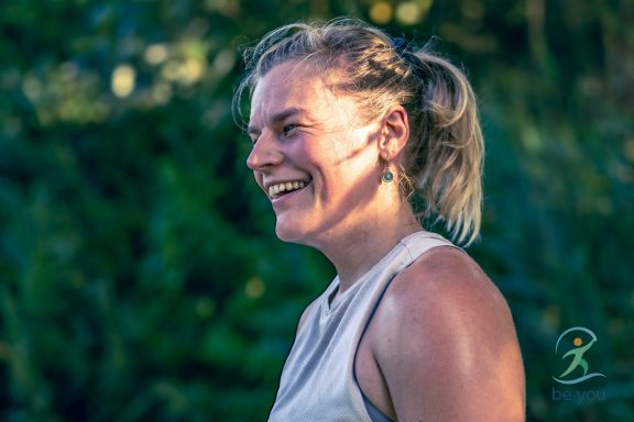 Lachende Frau mit kurzen blonden Haaren, umgeben von grüner Vegetation.