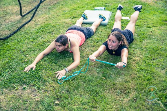 Zwei Frauen liegen auf dem Rasen und dehnen mit einem Gummiband.
