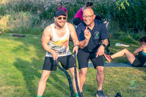 Zwei Männer trainieren im Freien mit einem Widerstandsband, einer gibt Anweisungen.