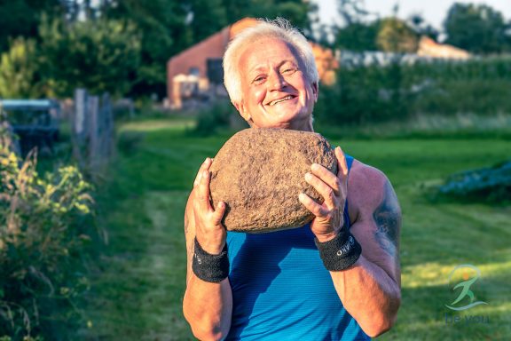 Älterer Mann in blauer Sportkleidung lächelt und hebt einen schweren Stein im Freien.
