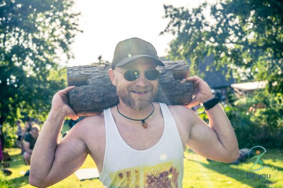 Mann mit Sonnenbrille und Hut, trägt einen Baumstamm auf den Schultern im Freien.