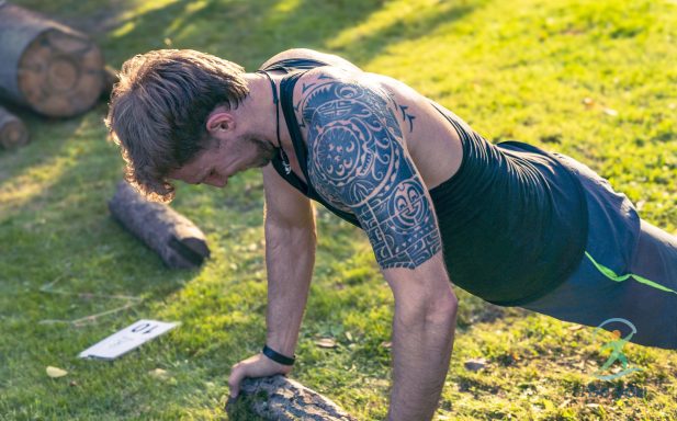 Mann macht Liegestütze im Freien auf Rasen, tätowierter Arm sichtbar.