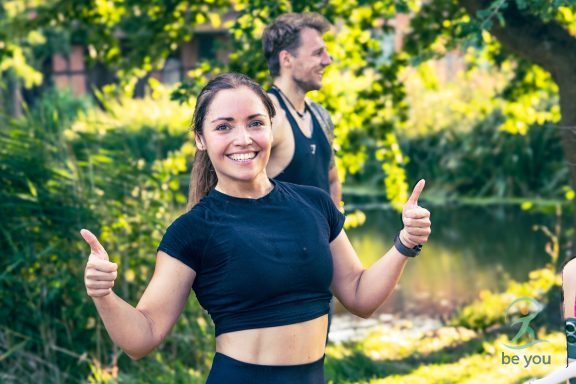 Junge Frau lächelt und zeigt Daumen hoch, im Hintergrund ein Mann und eine natürliche Umgebung.