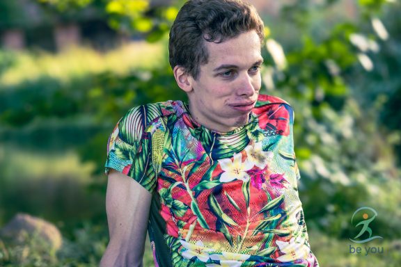 Junger Mann in bunter, floraler Kleidung sitzt im Freien vor grüner Kulisse.