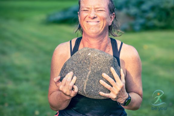 Frau hält einen großen Stein und lächelt mit Anstrengung auf einer Wiese.