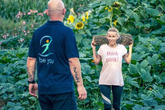 Eine Frau hebt einen Baumstamm, während ein Mann im Hintergrund zuschaut.