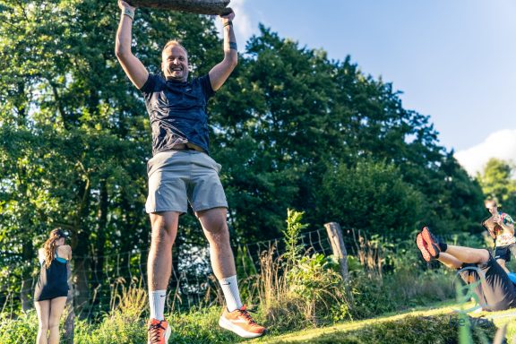 Mann hebt einen Baumstamm über Kopf, während andere im Hintergrund trainieren.