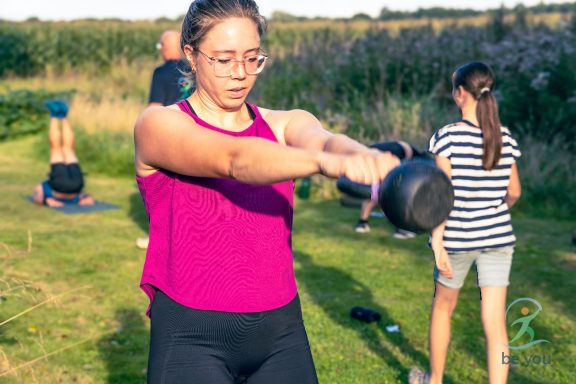 Frau mit einer Hantel, im Hintergrund andere Personen beim Training im Freien.