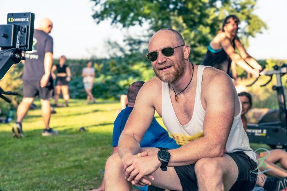 Lächelnder Mann mit Sonnenbrille sitzt im Freien, umgeben von Menschen bei sportlichen Aktivitäten.