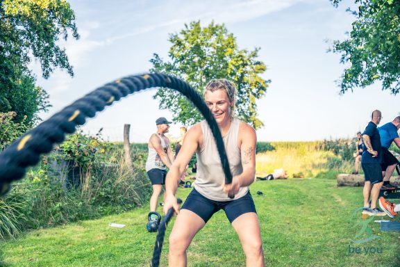 Eine Frau trainiert im Freien mit einem Battlerope. Im Hintergrund sind Bäume und Personen zu sehen.