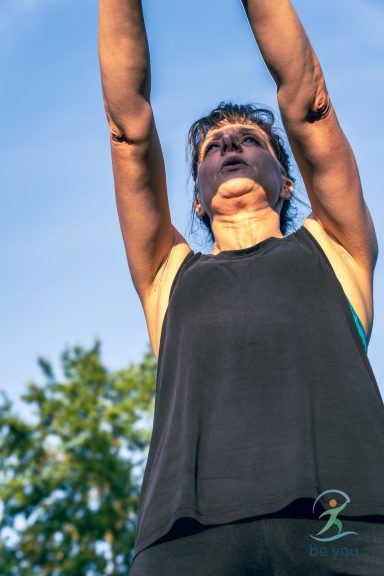 Frau mit hochgereckten Armen, im Freien, unter blauem Himmel.
