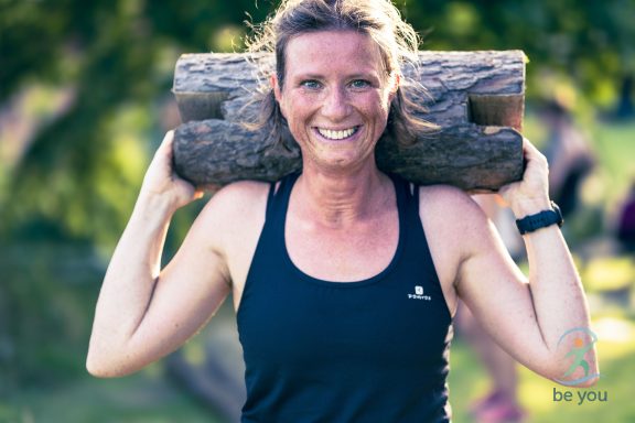 Frau mit Holzstamm auf den Schultern, lächelnd, in einem grünen Park.