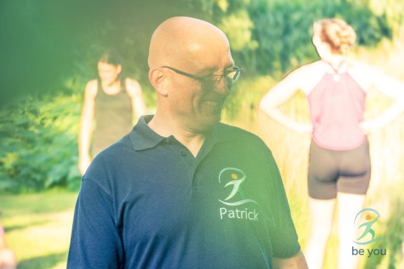 Mann in T-Shirt lächelt im Freien, im Hintergrund Frauen beim Sport.