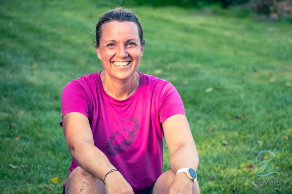 Lächelnde Frau mit kurzer Sportkleidung, sitzt lässig auf grünem Gras.