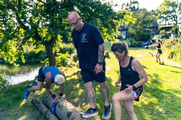Gruppentraining im Freien, Teilnehmer heben einen Baumstamm am Ufer eines Gewässers.