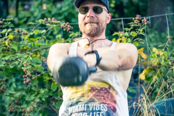 Mann mit Sonnenbrille, der eine Kettlebell im Freien hebt.
