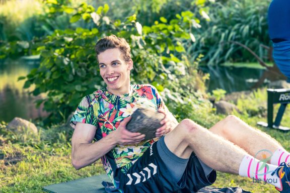 Junger Mann in buntem Shirt hält eine Kokosnuss und sitzt auf Wiese neben einem Gewässer.