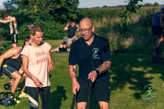 Zwei Personen beim Training im Freien, eine Frau steht neben einem Mann mit Trainingsgerät.