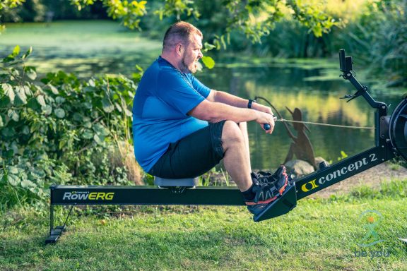 Mann beim Rudern auf einem Rudergerät im Freien, umgeben von Grün und Wasser.