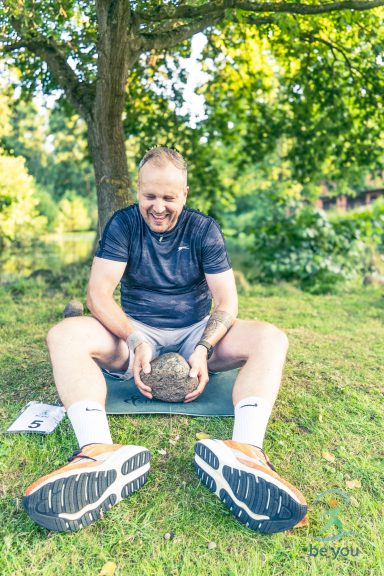Ein Mann sitzt im Freien und hält einen schweren Stein, umgeben von Grün.