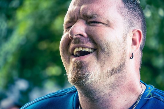 Lachender Mann mit kurzem Bart und Ohrpiercing, Hintergrund unscharf.