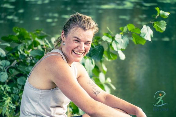 Junge Frau sitzt am Ufer, lächelt und umgeben von grünen Pflanzen.