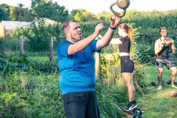 Mann in blauer T-Shirt hebt eine Kettlebell im Freien, Frauen im Hintergrund trainieren.