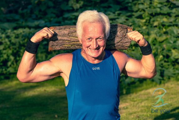 Älterer Mann hebt einen Baumstamm über den Schultern und lächelt.