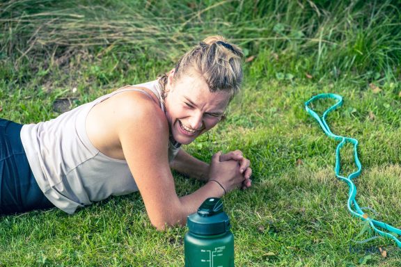 Eine lächelnde Frau liegt auf dem Gras neben einer Wasserflasche und einem grünen Kabel.