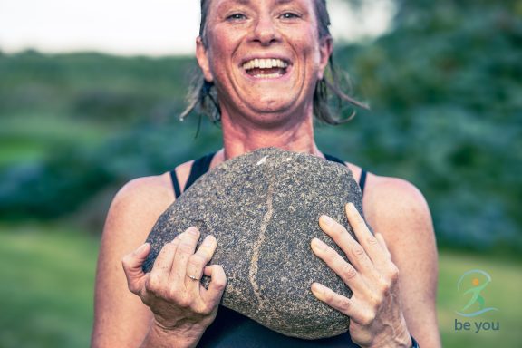 Lächelnde Frau hält einen großen Stein vor der Brust, umgeben von Natur.